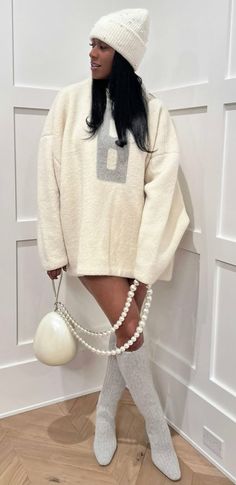 a woman wearing a white sweater and pearls is posing in front of a wall with her hands on her hips