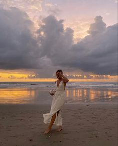 a woman standing on top of a sandy beach next to the ocean at sunset with clouds in the sky