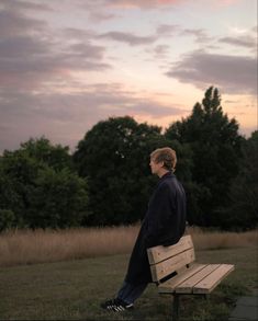 a man sitting on top of a wooden bench next to a field with trees in the background