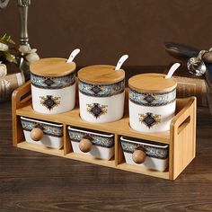 four ceramic containers with spoons and lids on a wooden tray in front of a vase