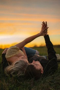 a man and woman laying in the grass with their hands up to each other as the sun sets behind them