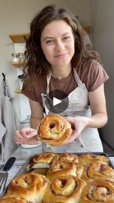 a woman holding up a pastry in front of a pan filled with cinnamon buns