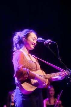 a woman holding a guitar while standing in front of a microphone and singing into a microphone