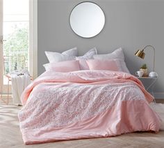 a bed with pink comforter and pillows in a white room next to a round mirror