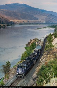 a train traveling down tracks next to a body of water with mountains in the background