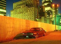 a red car parked next to a wooden fence in front of tall buildings at night