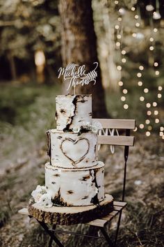 a white wedding cake sitting on top of a wooden bench in the woods with string lights behind it