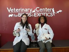 two women in white lab coats holding stuffed animals and posing for a photo with the veterinary center for birds and exotics sign behind them