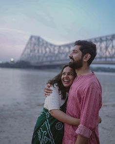 a man and woman standing next to each other near the water with a bridge in the background