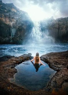 a woman sitting in the middle of a body of water surrounded by rocks and waterfalls