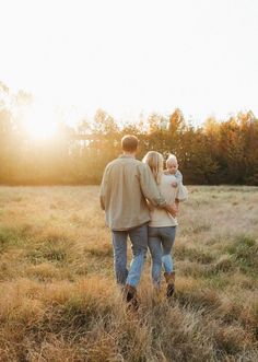 a man and woman are walking through the grass with their baby in his arms as the sun sets behind them