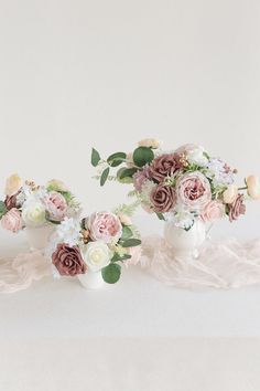 three white vases filled with pink and white flowers on top of a cloth covered table