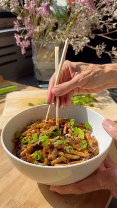 two hands holding chopsticks over a bowl of noodles with mushrooms and green onions