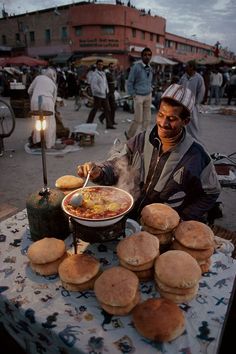 a man sitting at a table with food in front of him and people standing around