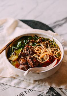 a white bowl filled with noodles, meat and vegetables next to chopsticks on top of a napkin