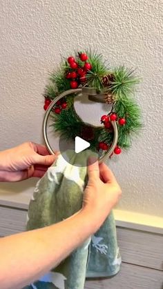 someone is holding up a christmas wreath on the wall above a towel rack with red berries and pine cones