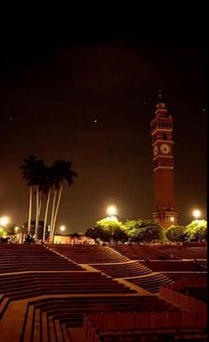 the clock tower is lit up at night in front of palm trees and benches with lights on them