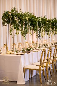 the table is set with white linens and gold chairs, greenery on each chair