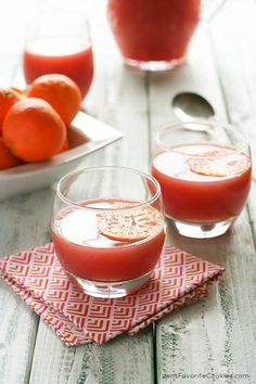 two glasses filled with red liquid sitting on top of a table next to oranges