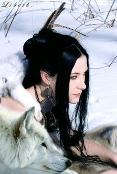 a woman with long black hair sitting in the snow next to two white wolfs