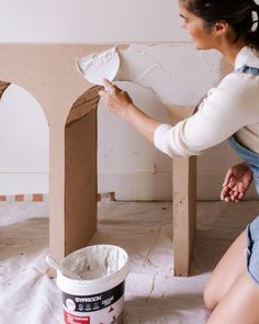 a woman kneeling down next to a bucket of white paint on the floor with a wooden structure in front of her