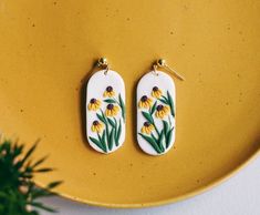 two yellow and white earrings with flowers painted on them sitting next to a potted plant