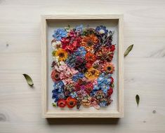 a wooden frame filled with colorful flowers on top of a white table next to green leaves