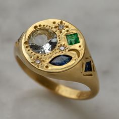 With its combination of colors and modern-meets-spiritual design, this Pari ring has a stunning sophistication. The 18K yellow gold round signet style ring face has one marquise blue sapphire, one round white sapphire and one square emerald on its front, along with three shimmering star set diamonds. Two additional blue sapphires are set within the 18K yellow gold shank on either side.