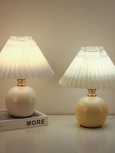 two white lamps sitting on top of a table next to a book and lamp shade