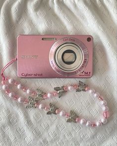 a pink camera sitting on top of a white bed next to a necklace and bracelet