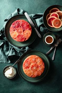 two plates topped with blood oranges and whipped cream next to silverware on a green surface