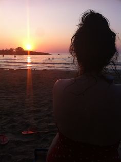 a woman sitting on the beach watching the sun go down over the ocean and people swimming in the water