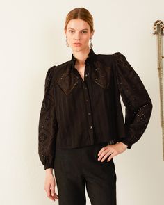 A person is dressed in the Annabel Ebony Eyelet Shirt, a black blouse with long sleeves that showcase intricate cutout patterns and a ruffled collar, complemented by decorative shoulder and arm details. The outfit is completed with high-waisted black pants, and the individual stands against a plain light-colored wall. Alix Of Bohemia, Eyelet Shirt, Ruffled Collar, French Seam, Long Puff Sleeves, Puffed Sleeves, Mother Of Pearl Buttons, If You Love, Unique Fashion