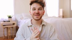 a man sitting in front of a bed with his hands folded up and looking at the camera