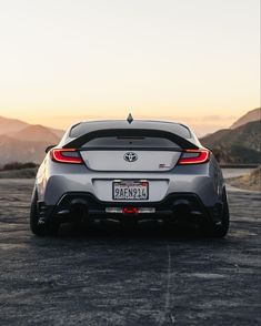 the rear end of a silver sports car parked in a parking lot at sunset with mountains in the background