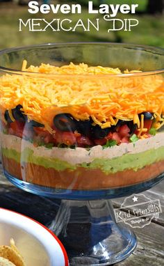 a layered mexican dip in a glass dish with chips on the side and text overlay that reads seven layer mexican dip