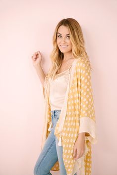 a woman standing in front of a pink wall wearing jeans and a yellow polka dot cardigan