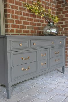 a gray dresser with gold handles and knobs on the drawers is sitting in front of a brick wall