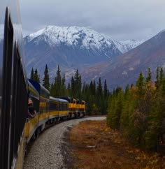 a train traveling through the mountains with snow covered mountains in the background and trees on either side