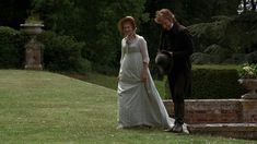 a man and woman dressed in period clothing walking through the grass with trees in the background