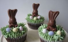 three cupcakes decorated with chocolate bunny ears and eggs in grass on a plate