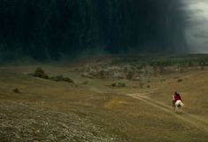 a person riding a horse down a dirt road in the middle of a field under a dark sky