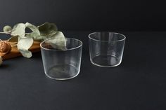 two clear cups sitting on top of a table next to a wooden stick and plant