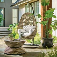 a wicker hanging chair with white pillows in front of a house and potted plants