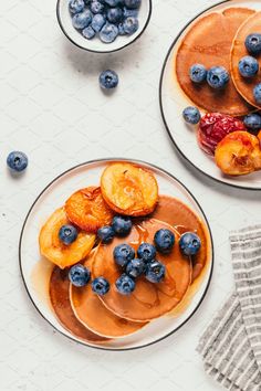 two plates filled with pancakes and blueberries on top of a white table next to a cup of coffee