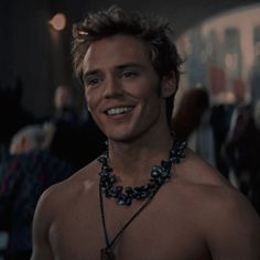 a shirtless man wearing a necklace and smiling at the camera with other people in the background