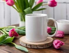 a white coffee cup sitting on top of a wooden table next to pink flowers and greenery