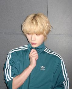 a young man with blonde hair wearing a green adidas jacket and holding his hand under his chin