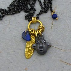 "A beautiful harmony of black oxidized silver chain , 22/24k solid gold, and deep blue Lapiz Lazuli comes together in this 24k gold pendants necklace. A cluster of several pendants hanging on a black silver chain, Lapis Lazuli bead and solid 24 karat gold leaf and a oxidized silver face. The chain is made of oxidized silver chain with 14k gold hoop claps. Dangle Lapis bead is attached to the claps. ~ 24k gold leaf pendant (with the bail): 0.2/0.75\" (5/19 mm). ~ Chain length : 20.5\" (52 cm). Th Blue Oxidized Necklace For Gift, Blue Oxidized Finish Necklace For Gift, Hand Forged Blue Pendant Necklace, Blue Hand Forged Pendant Necklace, Hand-forged Blue Pendant Necklace, Spiritual Blue Hand Forged Necklace, Gold Leaf Pendant, Silver Charm Necklace, Gold Hamsa