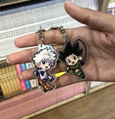 two anime keychains in the palm of someone's hand, with books behind them